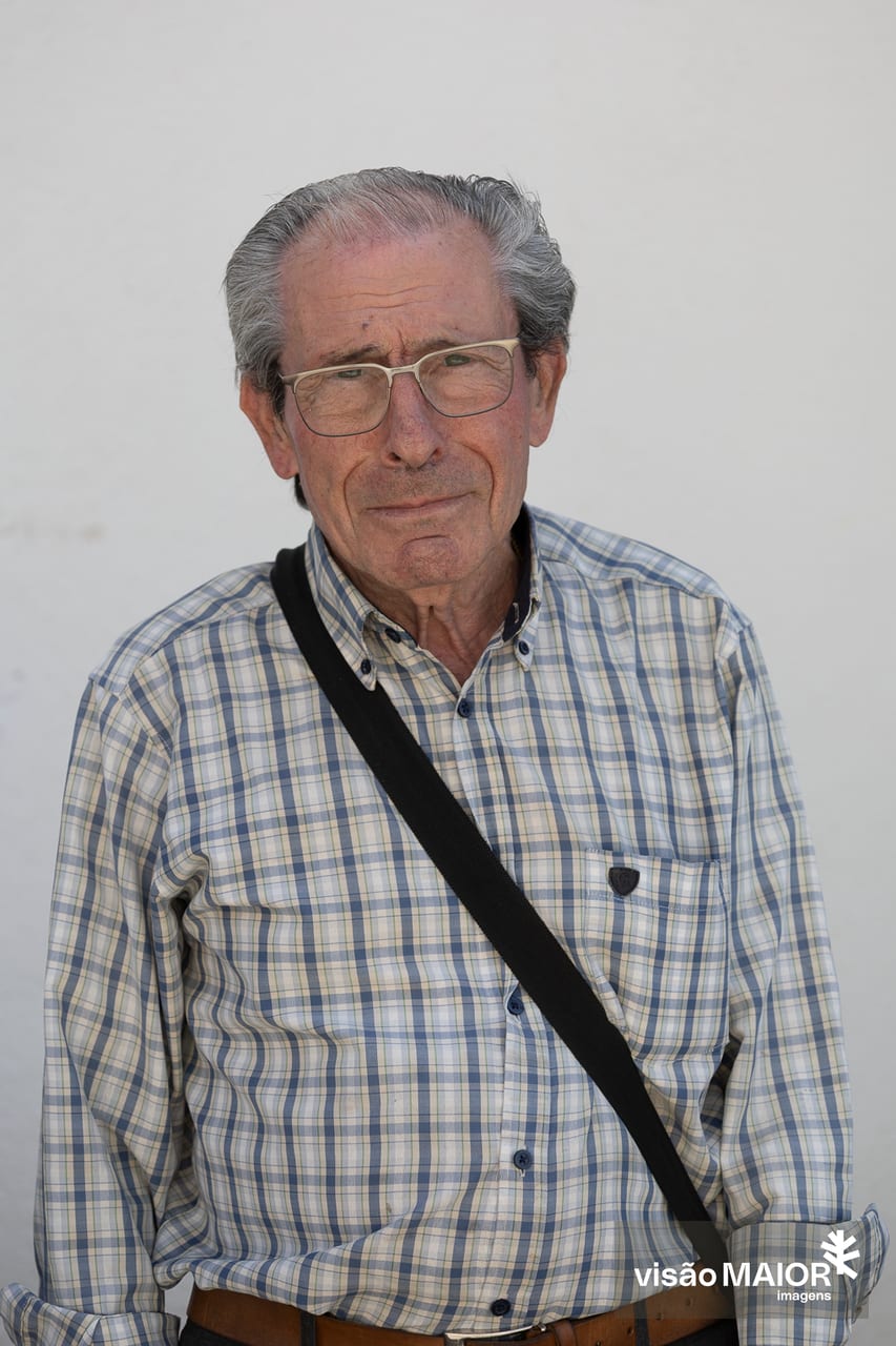Homem em pé, de cabelo branco, contemplativo de mala à tiracolo e camisa aos quadrados