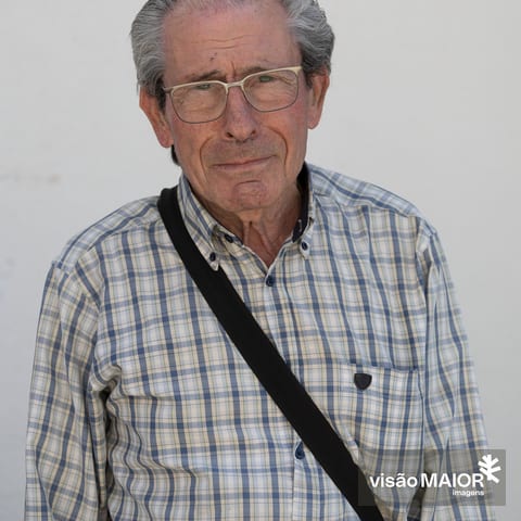 Homem em pé, de cabelo branco, contemplativo de mala à tiracolo e camisa aos quadrados