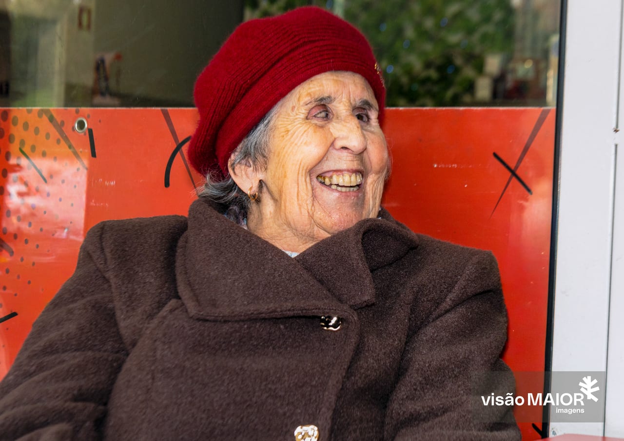Mulher de cabelo branco a olhar de lado, com gorro vermelho e sobretudo castanho a sorrir