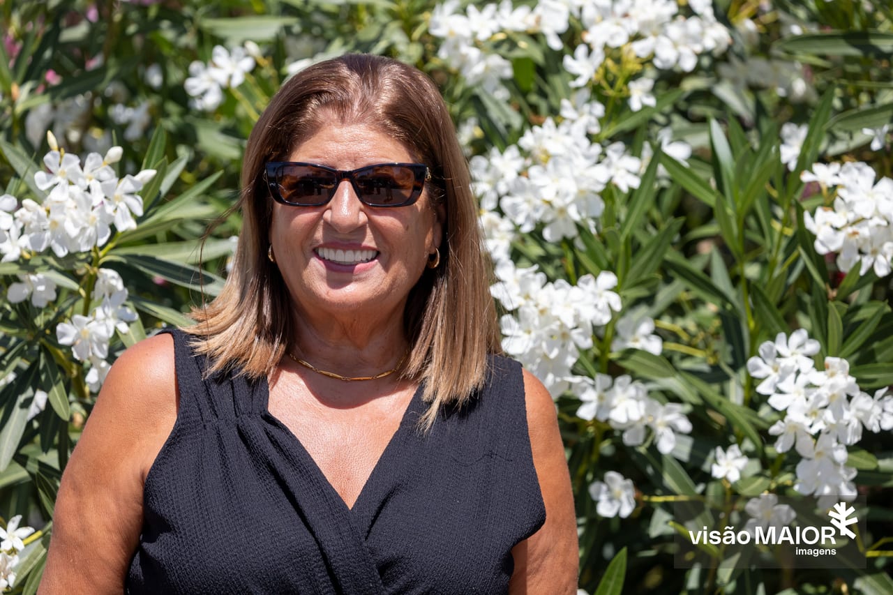 Mulher sorridente de cabelo castanho em pé, no meio de flores, de óculos escuros e top preto sem mangas