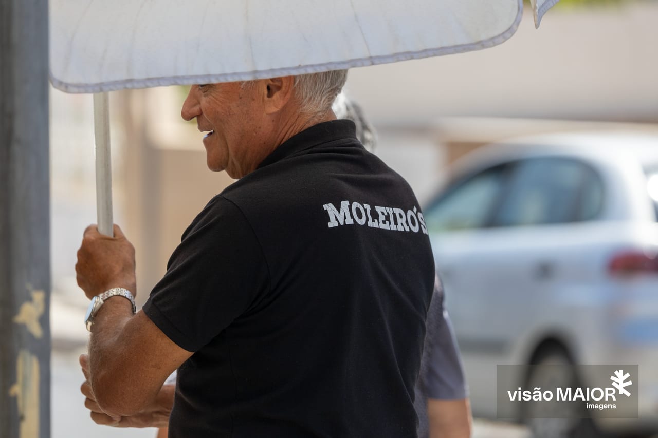 Homem de cabelo branco, de costas, a segurar um guarda sol, com t-shirt preta com um nome nas costas