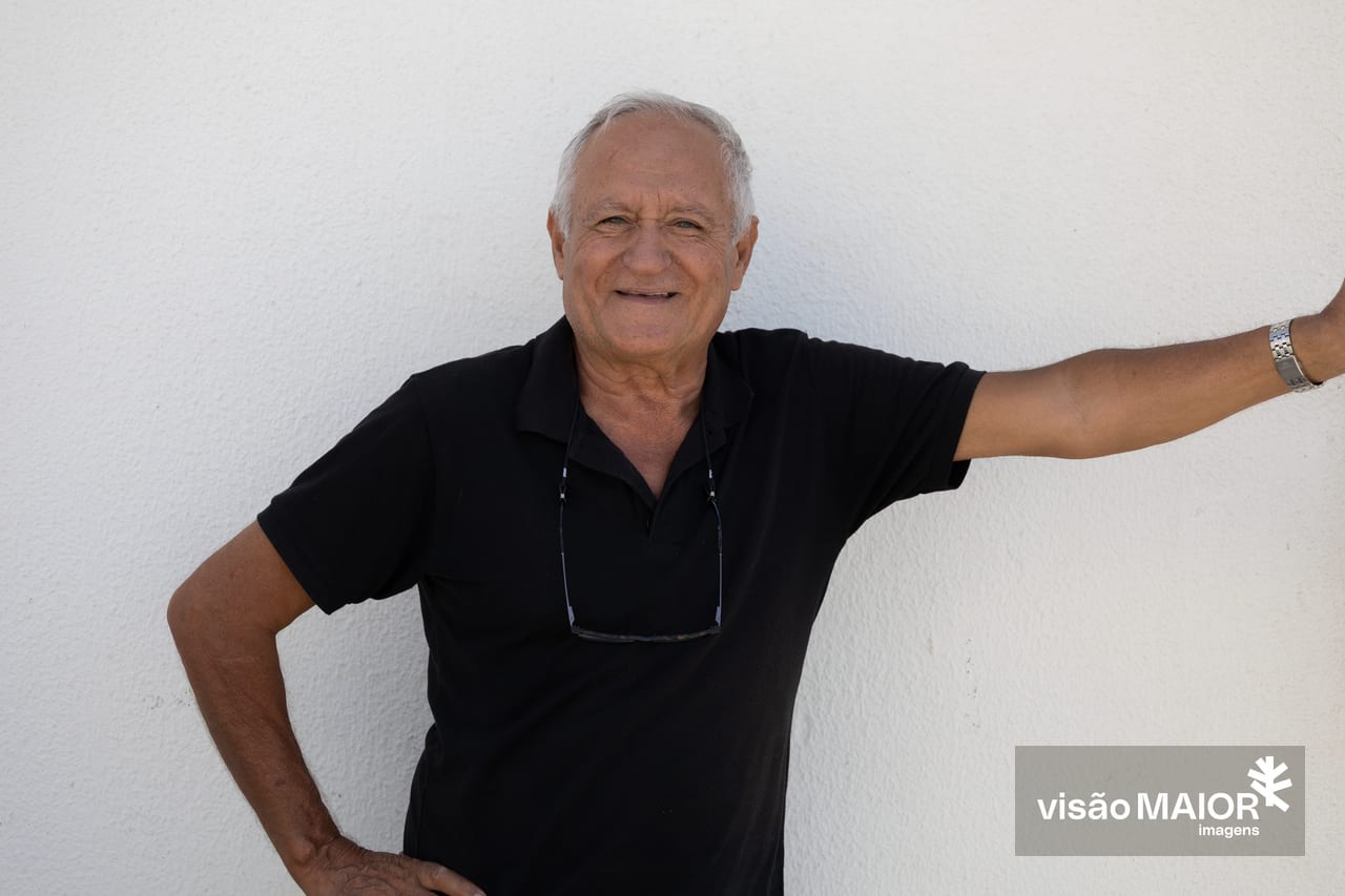 Homem de cabelos brancos e olhos claros, com os óculos caídos no peito, sorridente, com um braço na anca e o outro levantado
