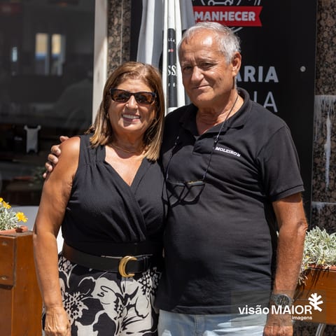 Casal abraçado em pé, ela de cabelos castanhos, ele de cabelos brancos, com um ar feliz e cumplice