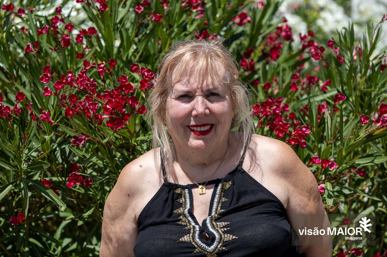 Mulher na rua, em frente a umas flores, cabelo claro e lábios pintados de vermelho, com top preto sem alças