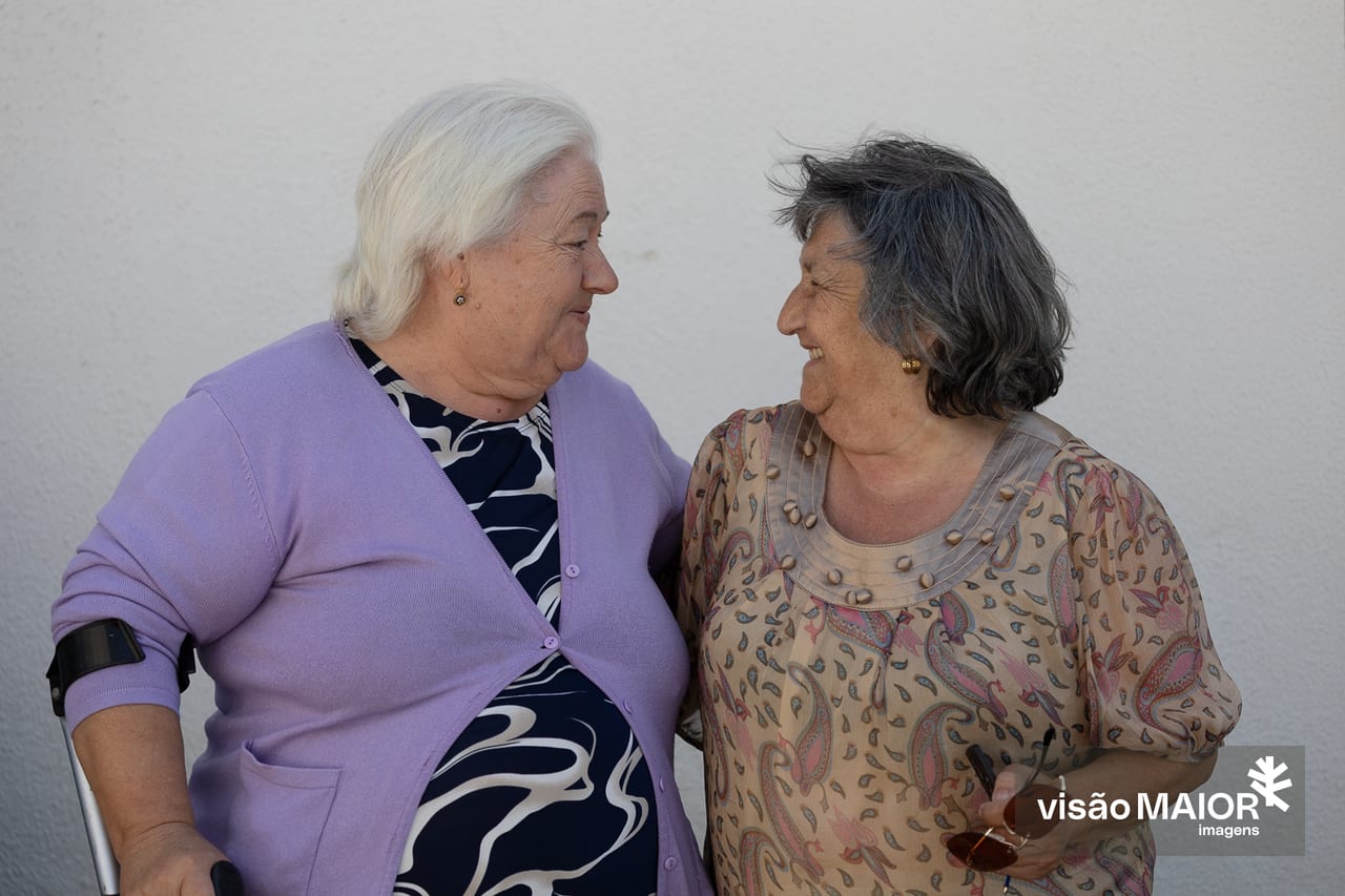 Duas mulheres de perfil, voltadas uma para a outra a rirem. Uma de cabelo preto, outra de cabelo branco