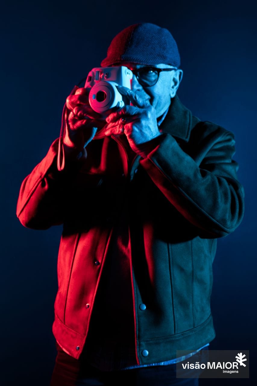 Homem de gorro, óculos e boné a tirar fotografias. Fundo escuro e reflexos vermelhos e azuis sobre ele.
