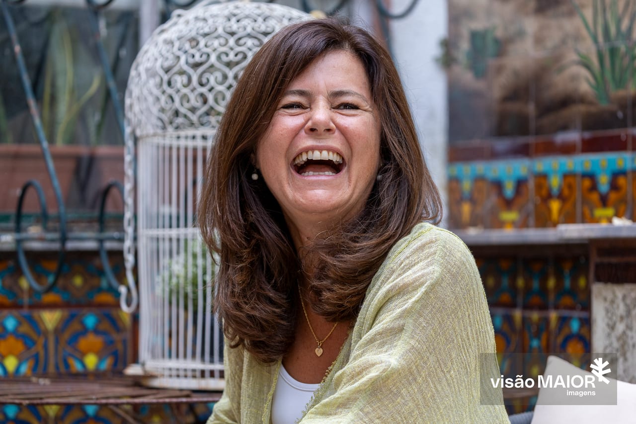 Mulher sentada, num páteo com azulejos, de colar e brincos a rir