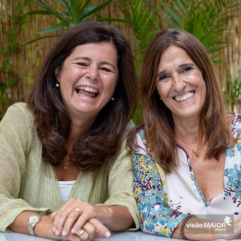 Duas mulheres ao ar livre, ambas de cabelo e olhos castanhos, com uma cerca de palhinhas e flores por trás a sorrirem