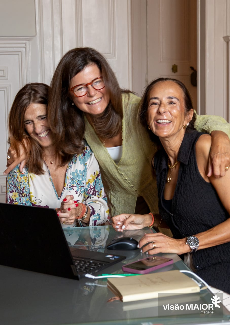 Três mulheres de cabelo castanho abraçadas, a do meio em pé e as outras sentadas, com um ar contente em frente a um computador