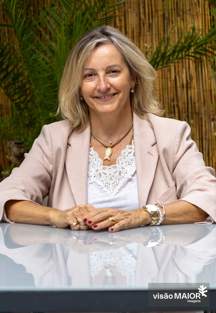 Mulher loira com roupa clara, com brincos, colar, relógio e pulseiras, sentada numa mesa ao ar livre com as mãos em cima mesa
