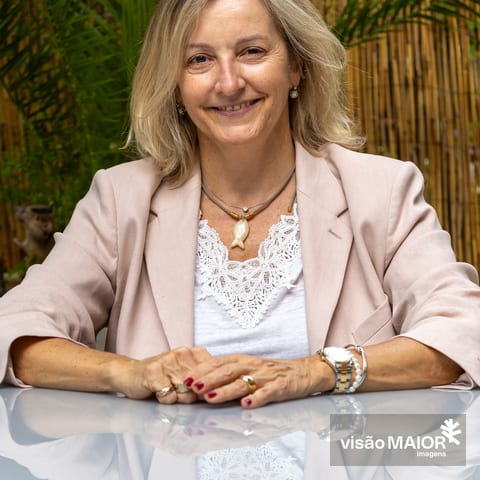 Mulher loira com roupa clara, com brincos, colar, relógio e pulseiras, sentada numa mesa ao ar livre com as mãos em cima mesa