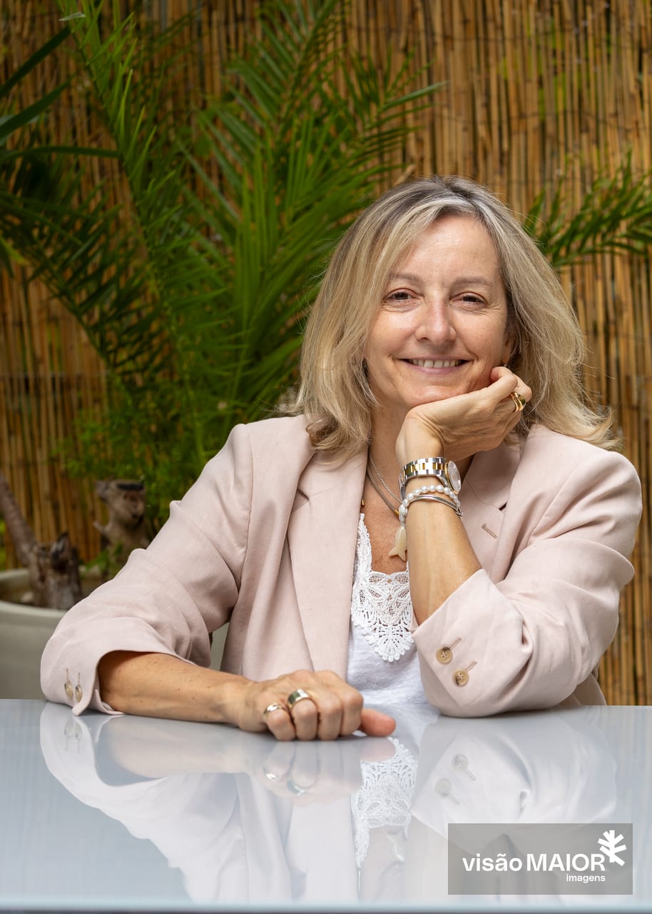 Mulher loira, com roupa clara sentada numa mesa ao ar livre com uma mão na mesa e outra a segurar o queixo