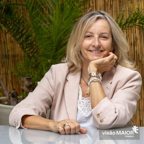 Mulher loira, com roupa clara sentada numa mesa ao ar livre com uma mão na mesa e outra a segurar o queixo