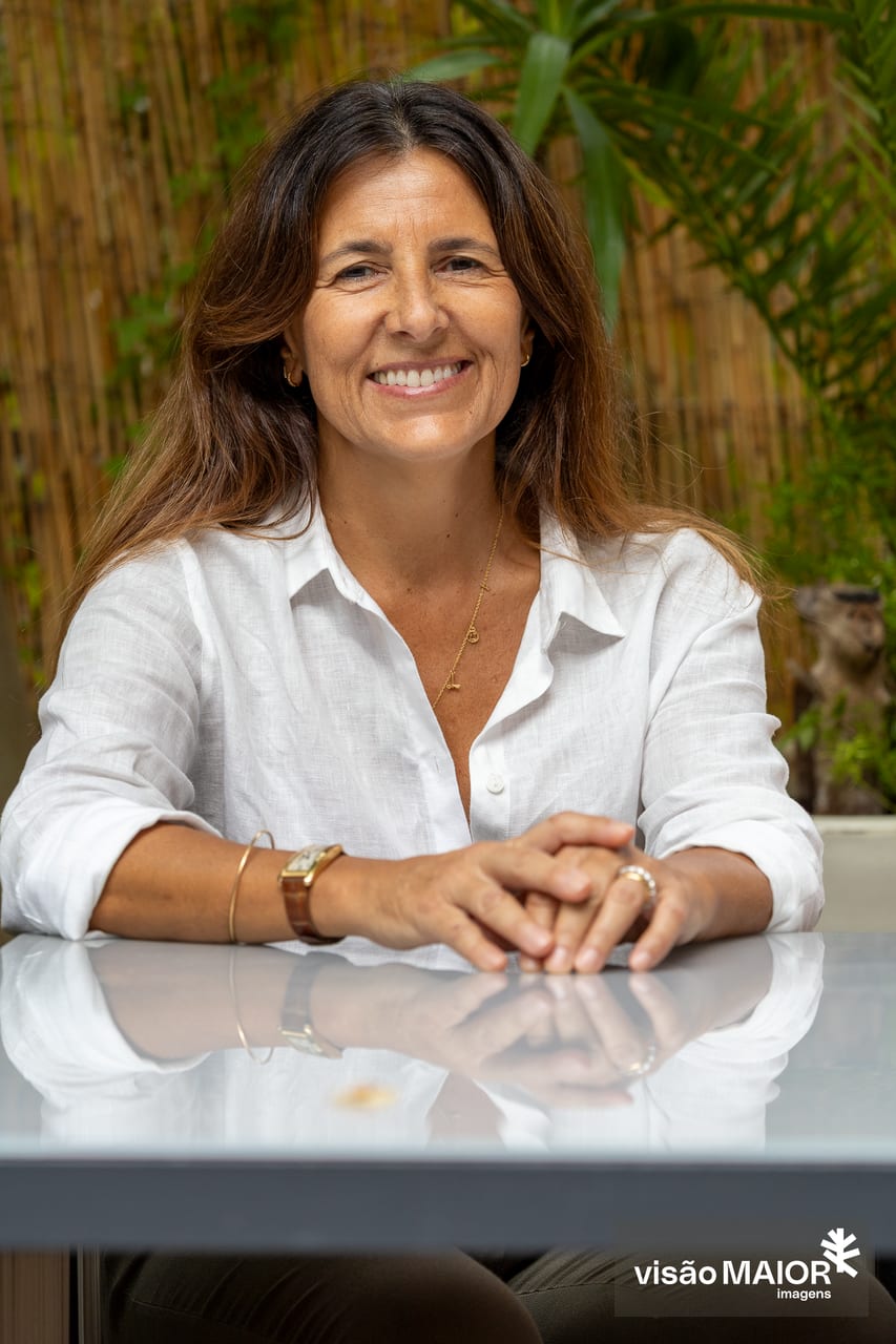 Mulher de cabelos castanhos a rir, com camisa branca, sentada em frente de uma mesa com os braços em cima, ao ar livre