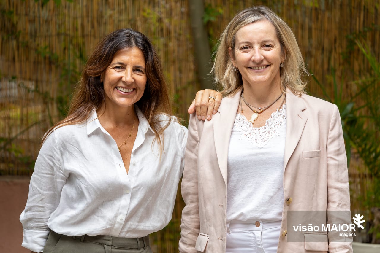 Duas mulheres em pé, uma com a mão no ombro da outra, uma loira, outra morena ao ar livre