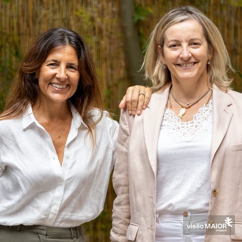 Duas mulheres em pé, uma com a mão no ombro da outra, uma loira, outra morena ao ar livre