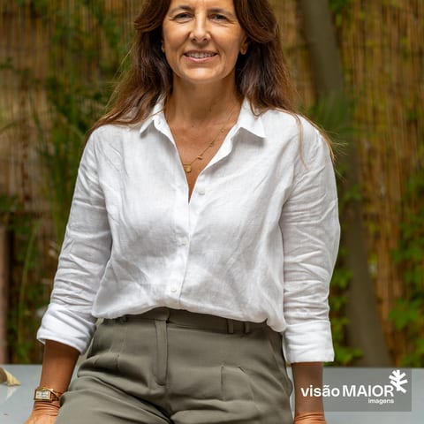 Mulher morena, sentada numa mesa, com camisa branca e calças verdes com as mãos apoiadas na mesa ao ar livre