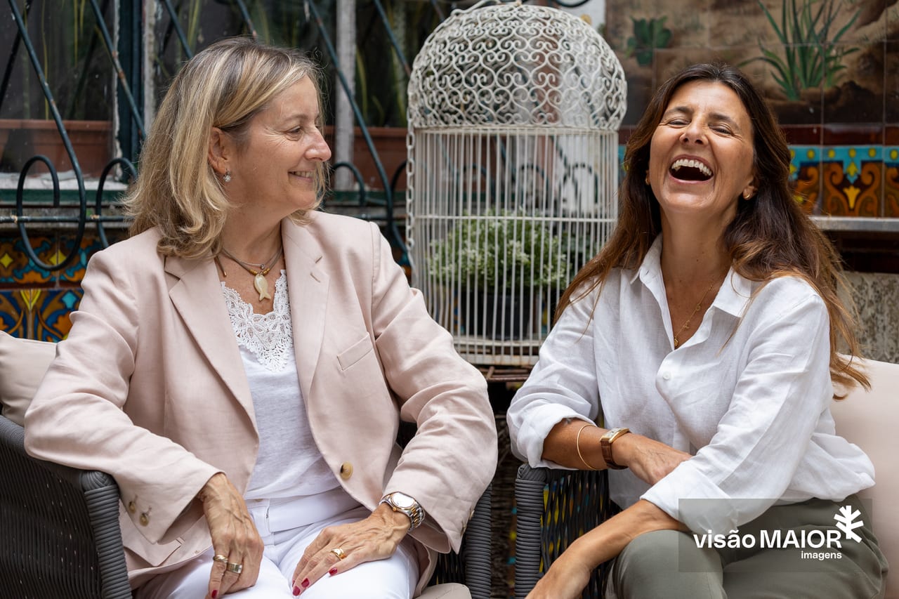 Duas mulheres, uma de cabelos loiros a olhar para uma de cabelos castanhos a rir, sentadas com uma gaiola por trás ao ar livre