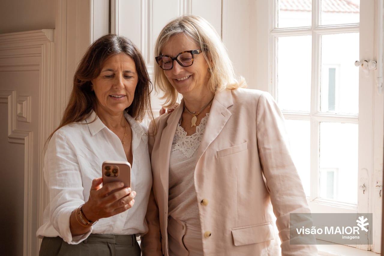 Duas mulheres em pé, uma morena de telemóvel na mão e outra loira de óculos a olhar para o telemovel