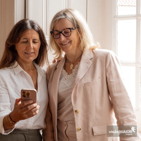 Duas mulheres em pé, uma morena de telemóvel na mão e outra loira de óculos a olhar para o telemovel