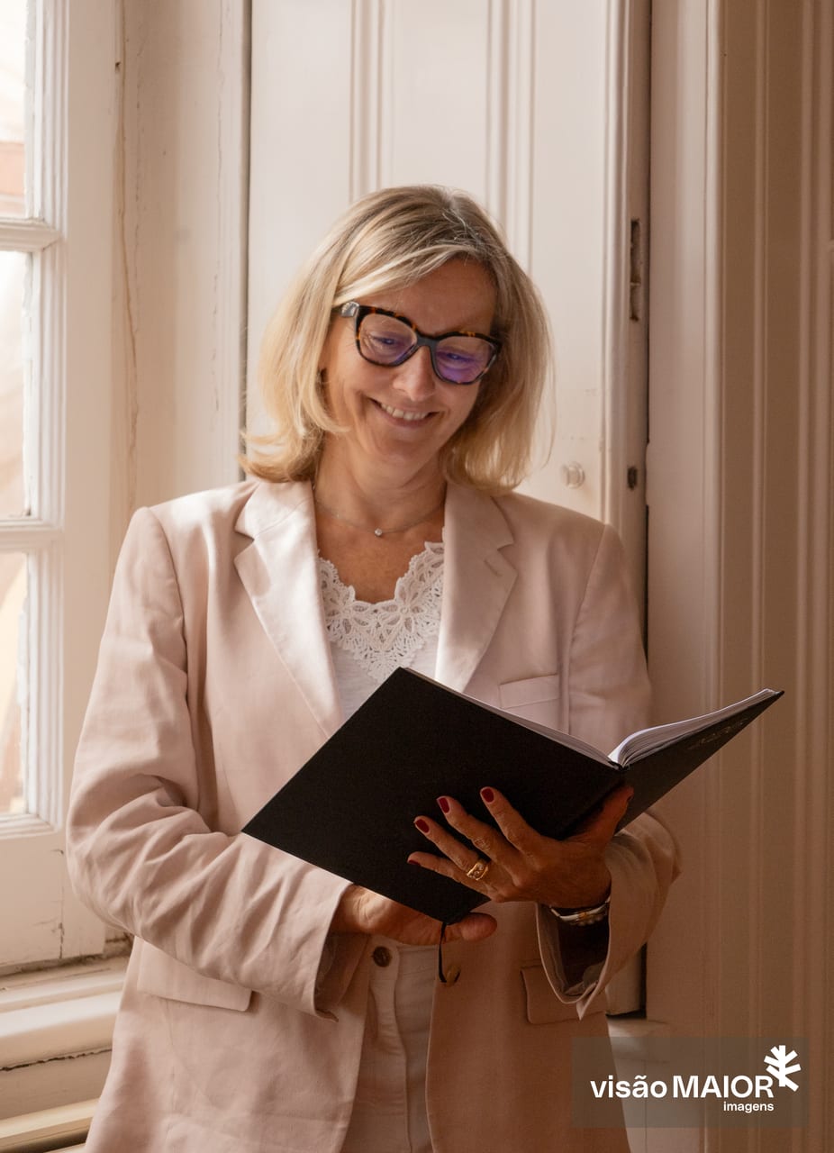 Mulher loira, com roupa clara e óculos em pé, a olhar para um livro e a sorrir